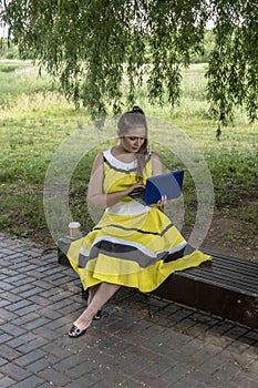 Young beautiful girl in a yellow dress with a laptop in the park. Works, learns, communicates online, making online shopping,