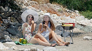 Young beautiful girls in hat and sunglasses drinking fresh coconut cocktails while talking and smiling on the sand beach