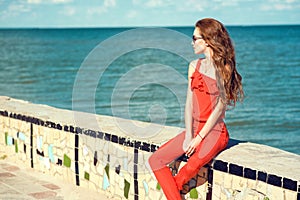 Young beautiful glam stylish woman wearing coral red jumpsuit and dark trendy sunglasses sitting on the parapet at the seaside