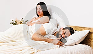 Young beautiful gorgeous sad girl sitting and hugging pillow on the bed in front of her boyfriend with mobile in hands.