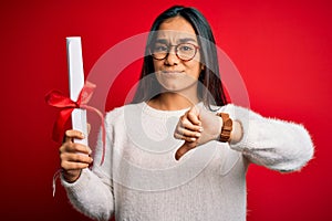 Young beautiful graduate asian woman holding university degree diploma over red background with angry face, negative sign showing