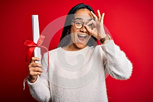 Young beautiful graduate asian woman holding university degree diploma over red background with happy face smiling doing ok sign