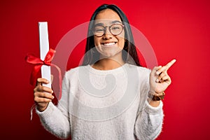 Young beautiful graduate asian woman holding university degree diploma over red background very happy pointing with hand and