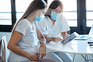 Young beautiful gynecologist wearing a hygienic face mask while showing to pregnant woman ultrasound scan baby with digital tablet