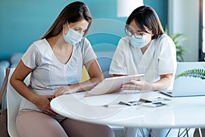 Young beautiful gynecologist wearing a hygienic face mask while showing to pregnant woman ultrasound scan baby with digital tablet