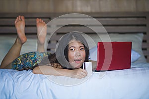 Young beautiful happy Asian American girl using credit card internet banking on laptop computer at home in bed shopping online e-c