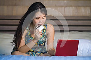 Young beautiful happy Asian American woman using credit card internet banking on laptop computer at home in bed shopping online e-