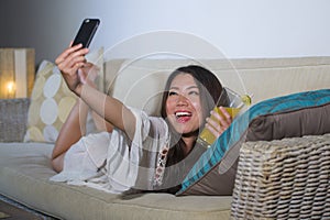 Young beautiful and happy Asian Chinese 20s or 30s woman taking selfie picture with mobile phone drinking orange juice at home liv