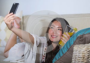 Young beautiful and happy Asian Chinese 20s or 30s woman taking selfie picture with mobile phone drinking orange juice at home liv