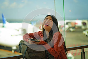 Young beautiful and happy Asian Chinese woman checking mobile phone holding passport at airport departure lounge carrying backpack