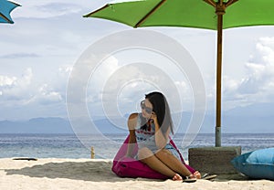 Young beautiful and happy Asian Chinese woman in chic Summer dress and sunglasses posing relaxed at tropical beach relaxed sitting