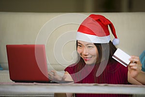 Young beautiful and happy Asian Chinese woman in Christmas Santa Claus hat holding credit card using laptop computer for internet