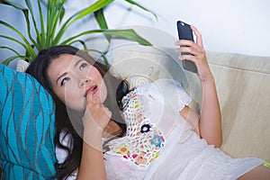 Young beautiful and happy Asian Chinese woman on her 20s or 30s lying at living room sofa couch using internet mobile phone lookin