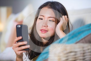 Young beautiful and happy Asian Chinese woman on her 20s or 30s lying at living room sofa couch using internet mobile phone lookin