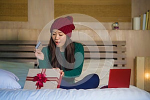 Young beautiful and happy Asian Chinese woman holding credit card and Christmas gift box shopping online xmas presents at home bed