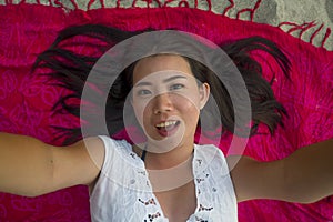 Young beautiful and happy Asian Chinese woman lying flat on sarong on beach sand smiling cheerful and relaxed taking selfie