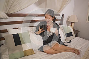 Young beautiful and happy Asian Chinese woman in nightgown drinking morning coffee relaxed in bed at home using mobile phone