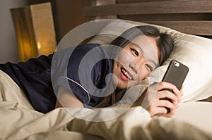 Young beautiful and happy Asian Chinese woman in pajamas using mobile phone social media texting with her boyfriend or enjoying