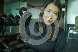 Young beautiful and happy Asian Chinese woman in sport hoodie posing cool at fitness center sitting by dumbbells rack during