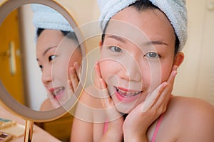 Young beautiful and happy Asian Chinese woman with towel head wrap applying makeup looking to mirror smiling cheerful preparing