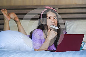 Young beautiful happy Asian Chinese woman using credit card internet banking on laptop computer at home in bed smiling shopping on