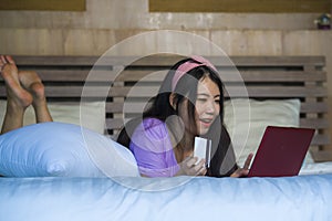 Young beautiful happy Asian Chinese woman using credit card internet banking on laptop computer at home in bed smiling shopping on