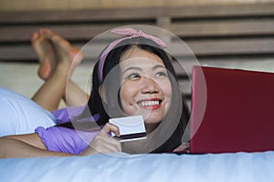 Young beautiful happy Asian Chinese woman using credit card internet banking on laptop computer at home in bed smiling shopping on