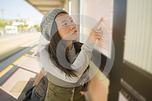 Young beautiful and happy Asian Chinese woman waiting the train on station platform bench checking travel and itinerary schedule