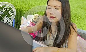 Young beautiful and happy Asian Chinese woman working outdoors with laptop computer networking green grass background cafe as digi