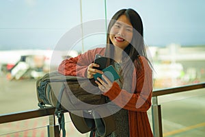 Young beautiful and happy Asian Japanese woman checking mobile phone holding passport at airport departure lounge carrying