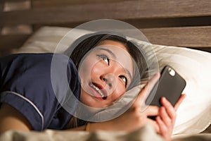 Young beautiful and happy Asian Japanese woman in pajamas using mobile phone social media texting with her boyfriend or enjoying