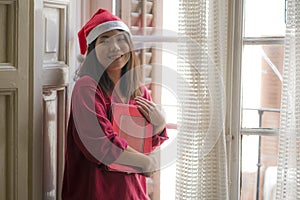 Young beautiful and happy Asian Japanese woman in Santa Claus hat with Christmas present at home feeling cheerful and positive