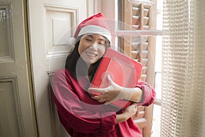 Young beautiful and happy Asian Japanese woman in Santa Claus hat with Christmas present at home feeling cheerful and positive