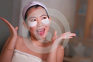 Young beautiful and happy Asian Japanese woman smiling to bathroom mirror applying hydrating eye patches under the eyes in beauty