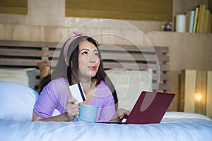 Young beautiful happy Asian Japanese woman using credit card internet banking on laptop computer at home in bed shopping online e-