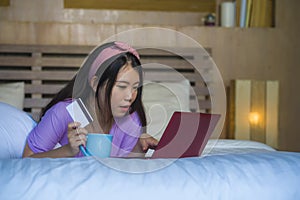 Young beautiful happy Asian Japanese woman using credit card internet banking on laptop computer at home in bed shopping online e-