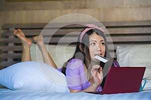 Young beautiful happy Asian Japanese woman using credit card internet banking on laptop computer at home in bed smiling shopping o