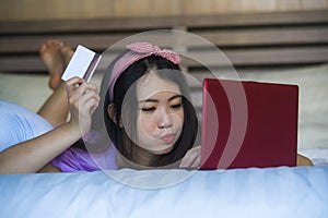 Young beautiful happy Asian Japanese woman using credit card internet banking on laptop computer at home in bed smiling shopping o