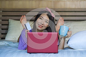 Young beautiful happy Asian Japanese woman using credit card internet banking on laptop computer at home in bed smiling shopping o
