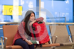 Young beautiful and happy Asian Korean tourist woman with travel suitcase in airport sitting at departures lounge gate waiting for