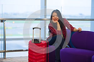 Young beautiful and happy Asian Korean tourist woman with travel suitcase in airport sitting at departures lounge gate waiting for