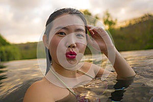 Young beautiful and happy Asian Korean woman in bikini enjoying sunset taking selfie at amazing jungle resort infinity pool