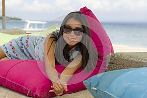 Young beautiful and happy Asian Korean woman in chic Summer dress and sunglasses posing relaxed at tropical beach relaxed sitting