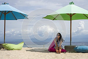 Young beautiful and happy Asian Korean woman in chic Summer dress and sunglasses posing relaxed at tropical beach relaxed sitting