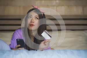 Young beautiful and happy Asian Korean woman at home bedroom using credit card internet banking with mobile phone in bed smiling o