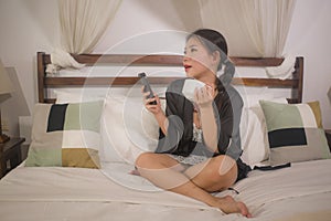 Young beautiful and happy Asian Korean woman in nightgown drinking morning coffee relaxed in bed at home using mobile phone