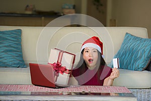 Young beautiful and happy Asian Korean woman in Santa hat holding credit card and Christmas present box using laptop computer for