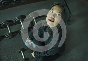 Young beautiful and happy Asian Korean woman in sport hoodie posing cool at fitness center sitting by dumbbells rack during