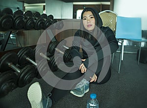 Young beautiful and happy Asian Korean woman in sport hoodie posing cool at fitness center sitting by dumbbells rack during