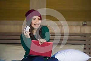 Young beautiful happy Asian Korean woman in winter hat relaxed on bed holding credit card using laptop computer for internet bank